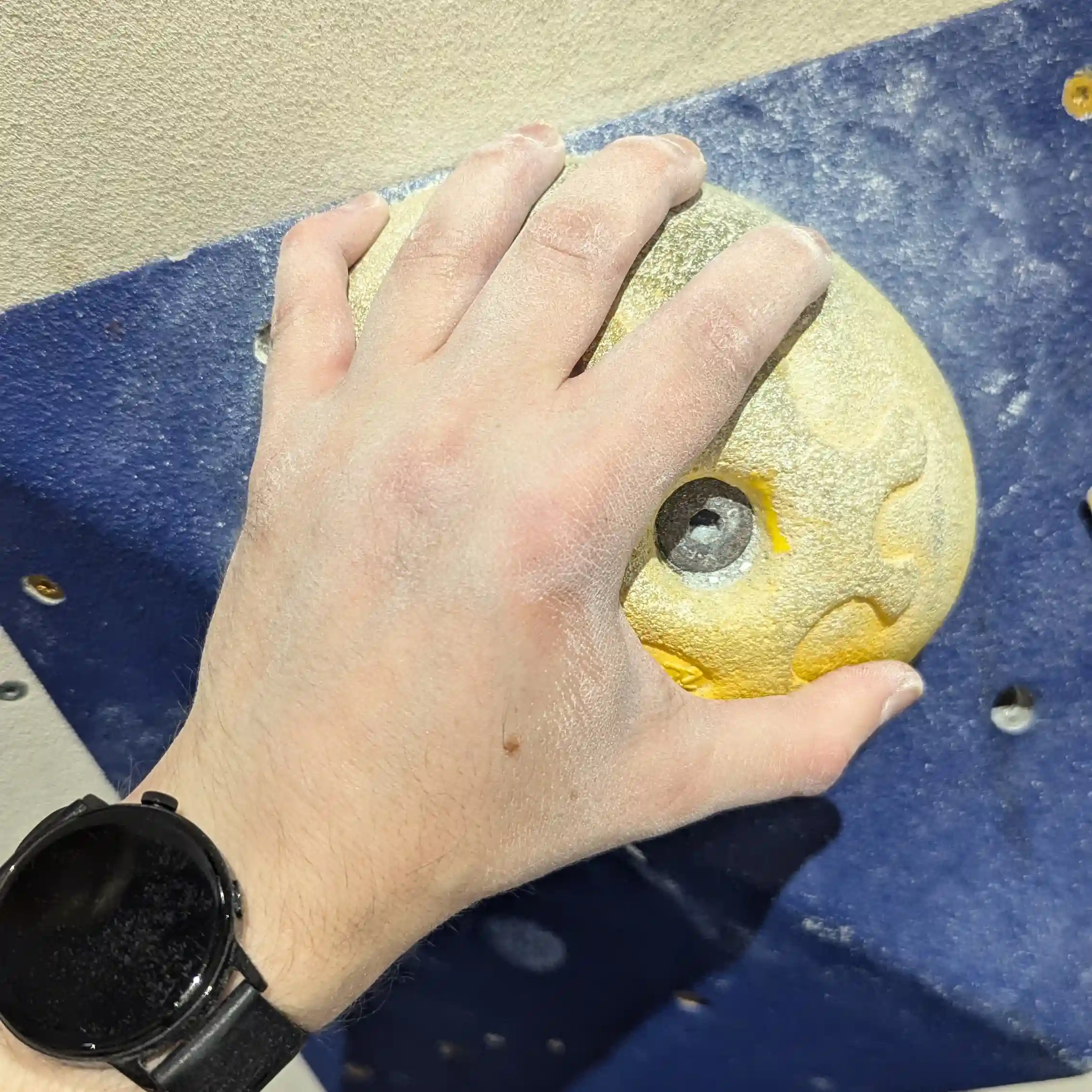 Shows a climbers left hand gripping the yellow, melon shaped hold with their full hand.