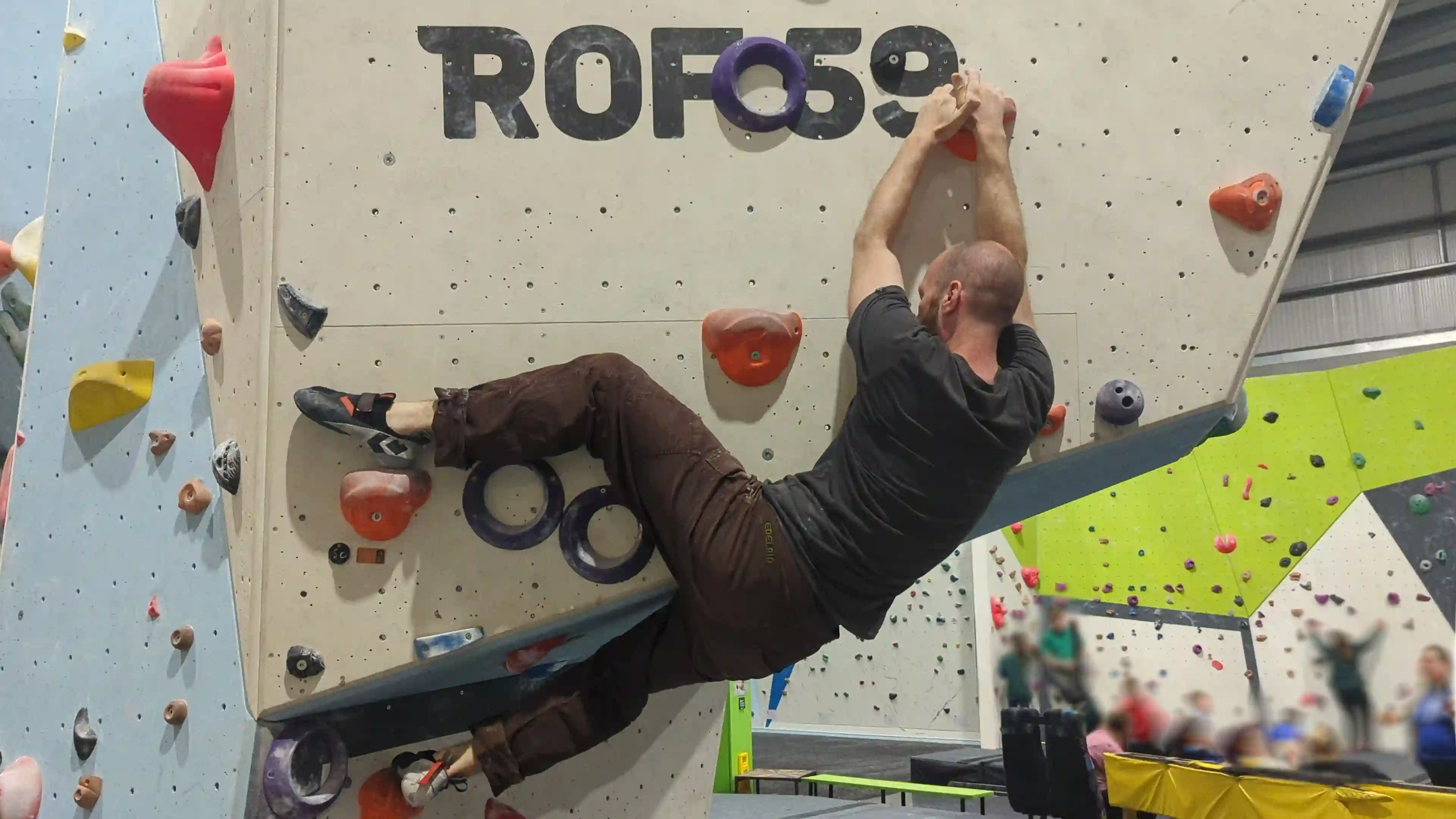 Shows the climber on the bouldering wall using the heel hook technique to help support their weight while gripping hte next hold.