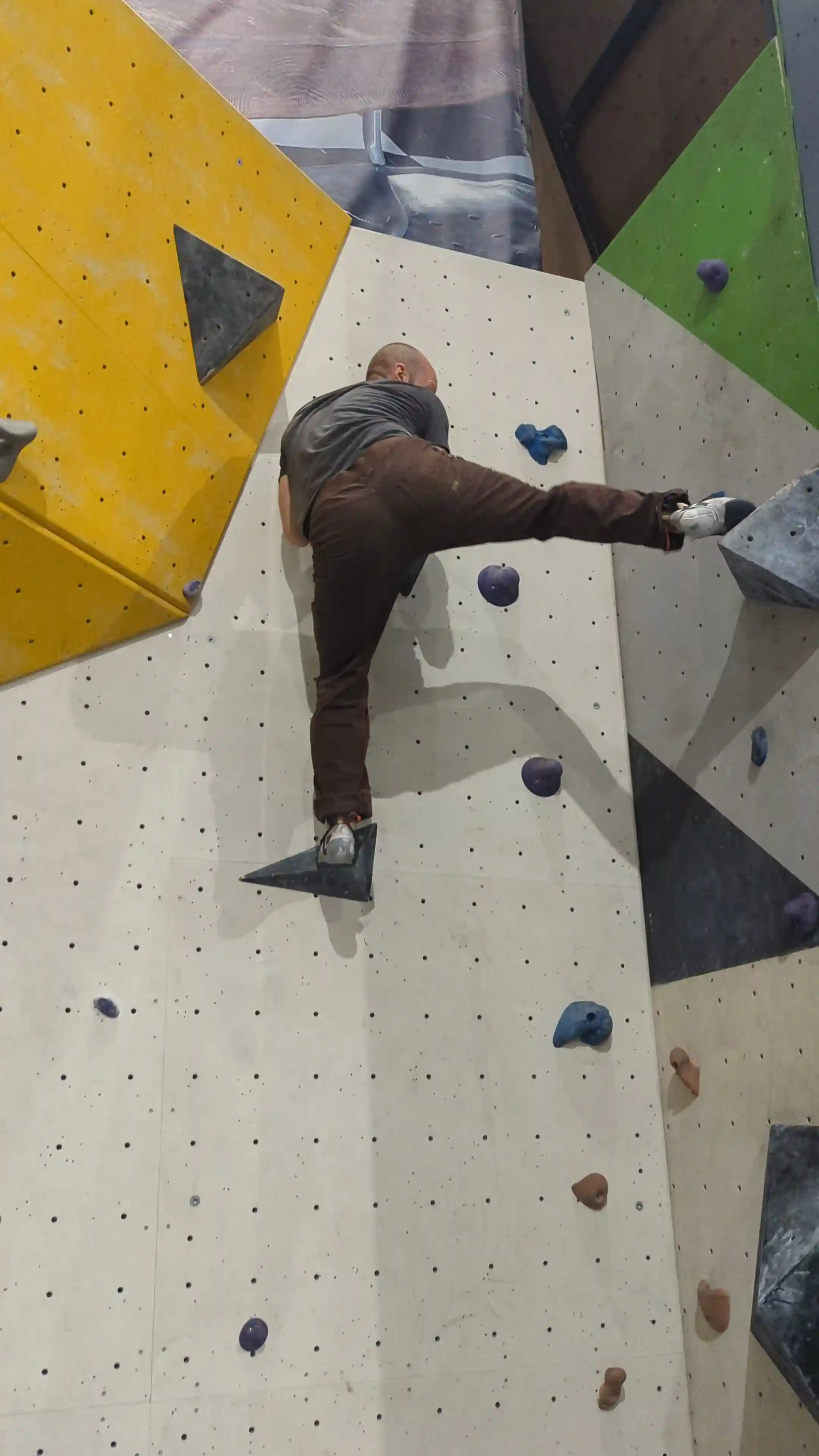 Shows the climber on the bouldering wall just before final hold. With right leg fully extend out to the right and the ball of their foot resting on a large triangular volume.