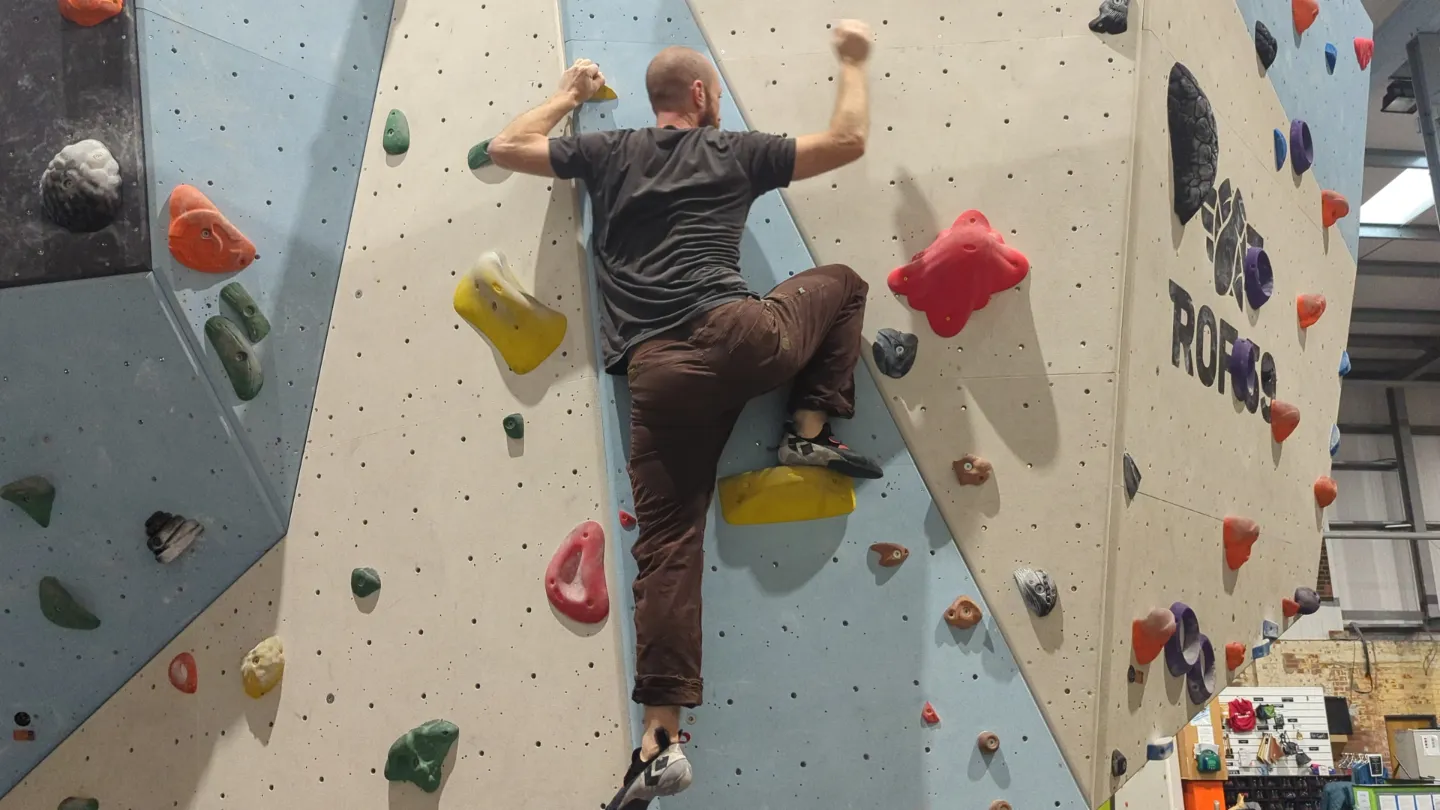 Shows the climber on the bouldering wall with their right leg far over on the foothold in the rock position, left leg hanging straight down and arm up above head to maintain balance.