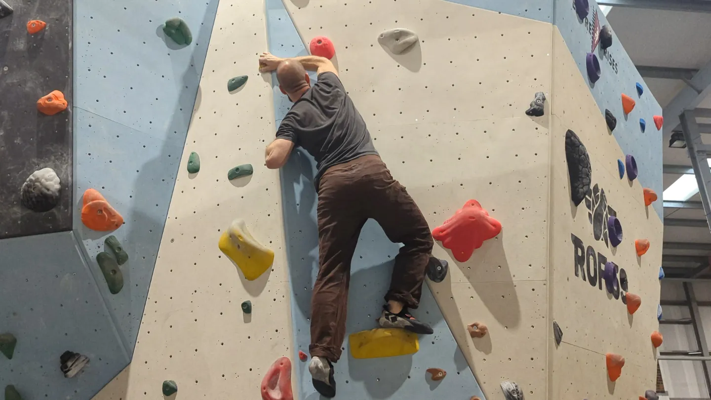 Shows the climber on the bouldering wall with almost fully extended right leg and moving their hands onto the next hold above them.