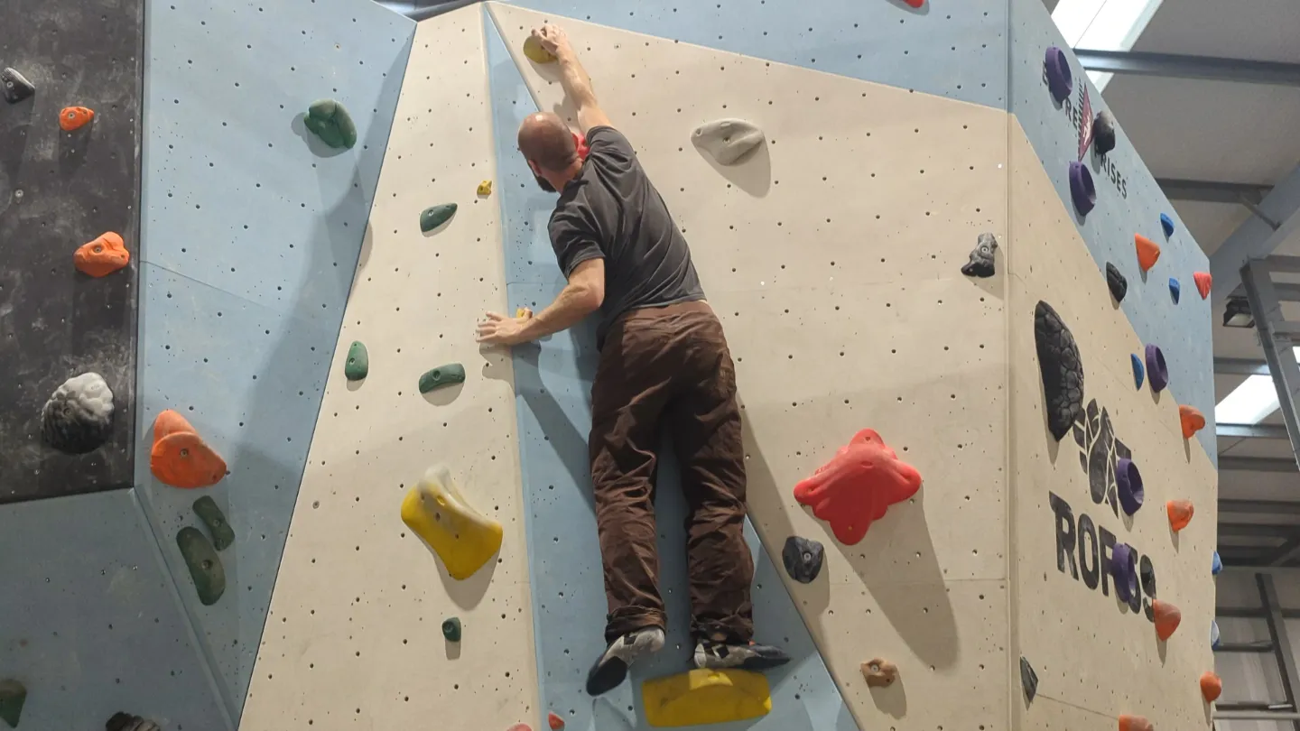 Shows the climber on the bouldering wall in the position to reach the top hold. Right leg is fully extended and center of gravity shifted to over both feet.