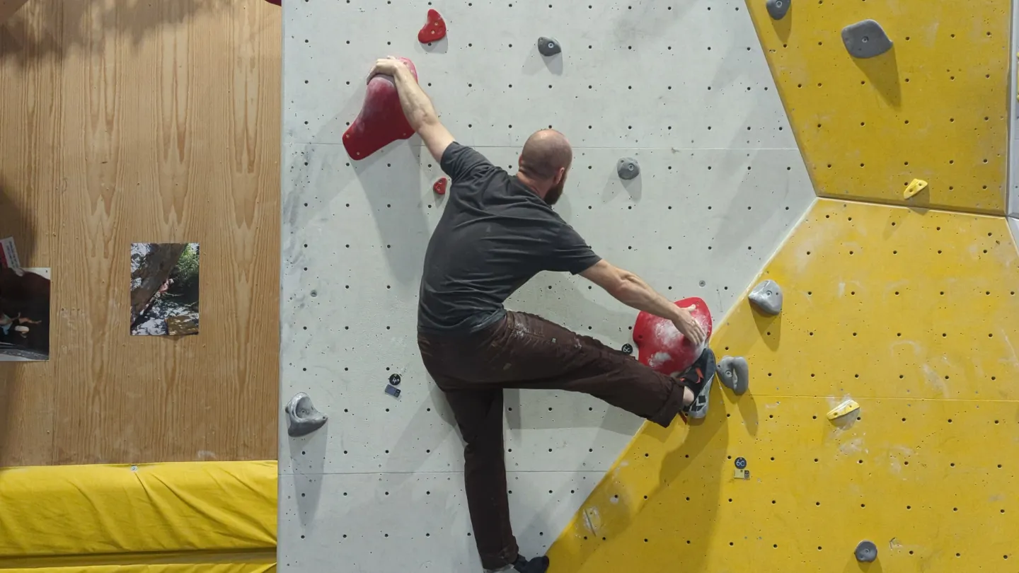 Shows the climber on the bouldering wall with his toe in position hooked into a large red hold.